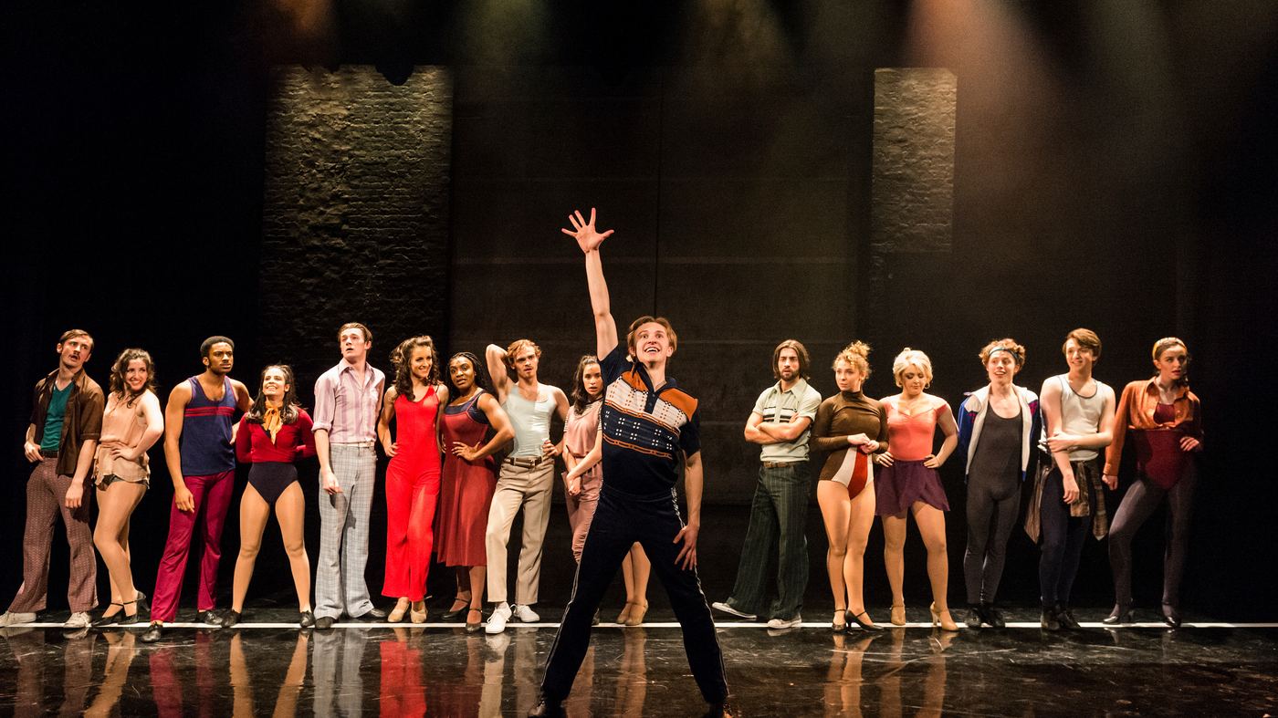 A full cast lined up on stage under theatre lighting